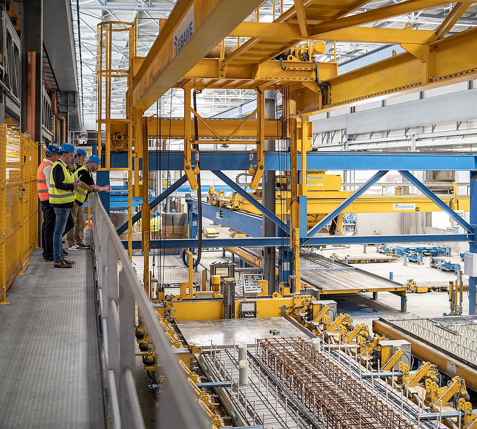 Mitarbeiter mit Schutzhelmen beobachten eine EBAWE-Fertigungsanlage zur Betonfertigteilproduktion in einer Werkshalle